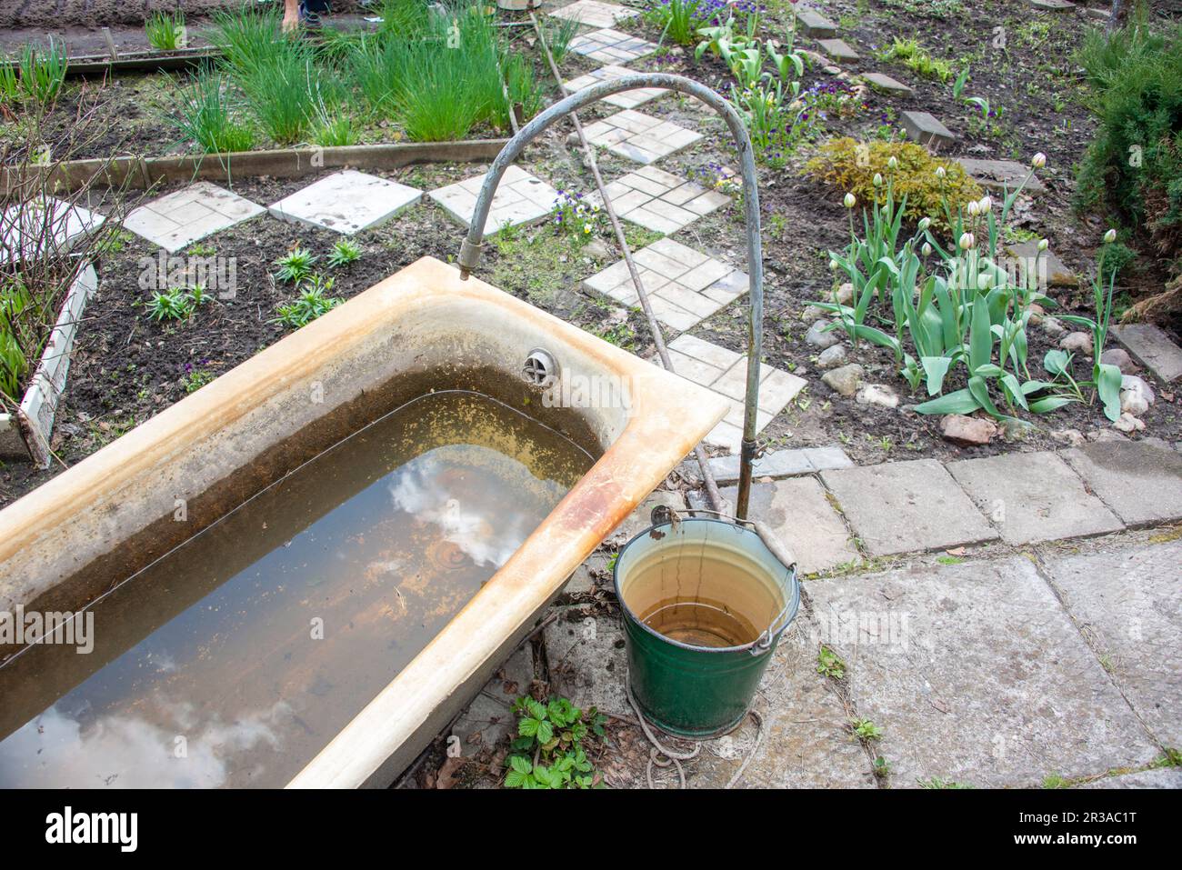 Vecchia vasca in ghisa con acqua, un secchio e un rubinetto per innaffiare le piante in giardino. Giardino, rurale, cottage. Foto Stock
