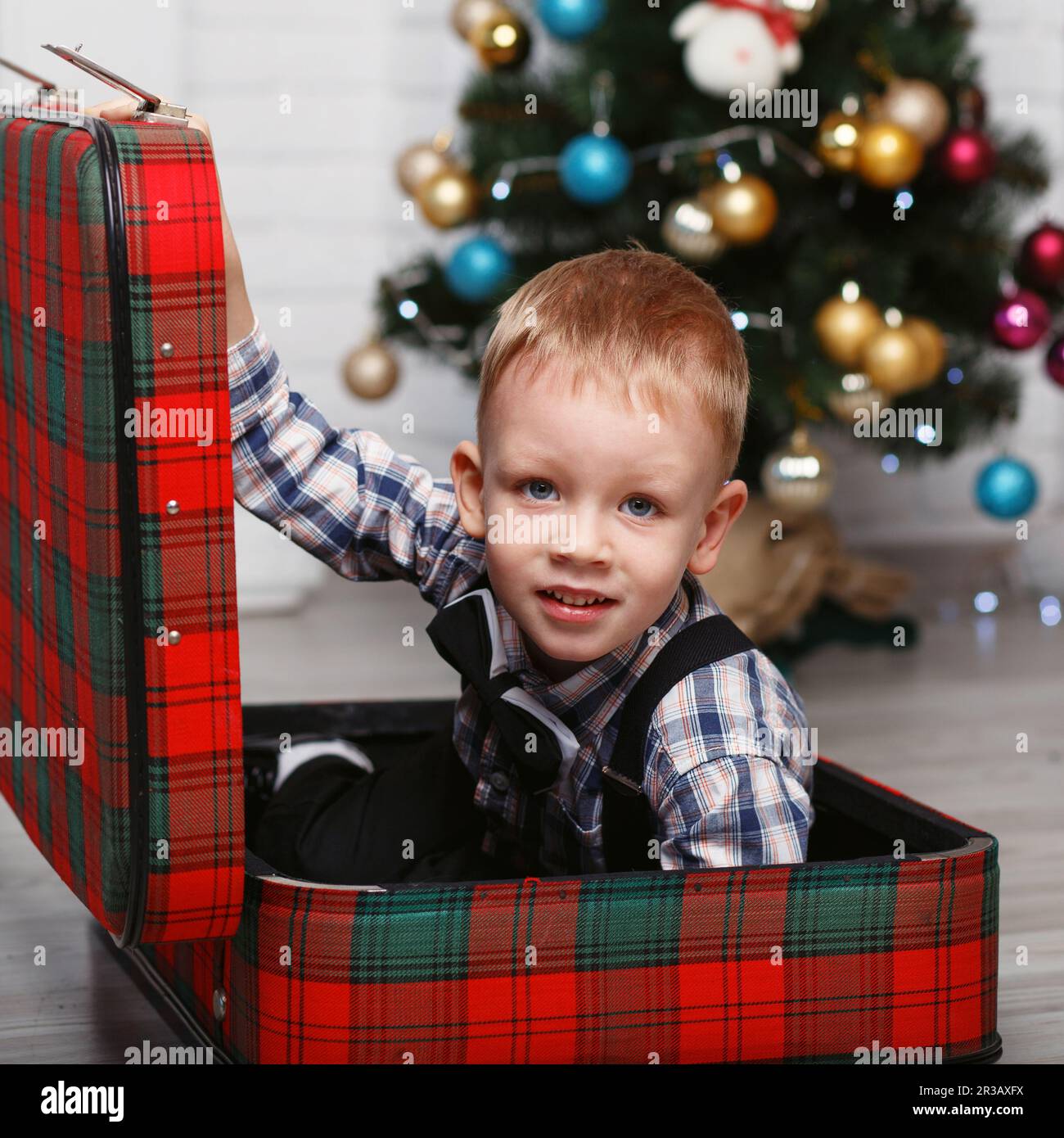 Carino ragazzo che gioca nascondendosi in una valigia rossa plaid Nell'interno con decorazioni natalizie Foto Stock