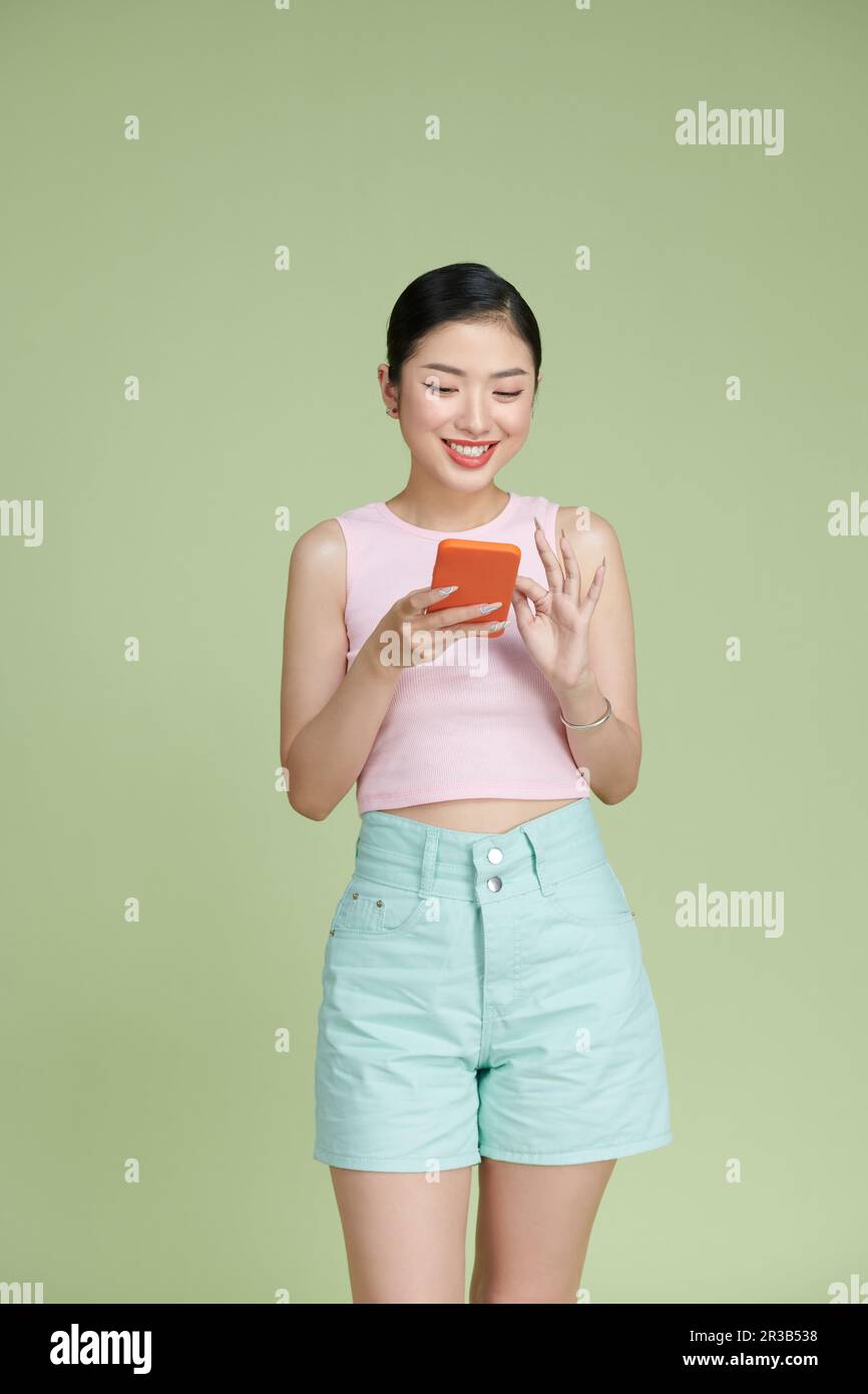 Sorridente affascinante donna asiatica utilizza cellulare felice SMS isolato su sfondo verde Foto Stock