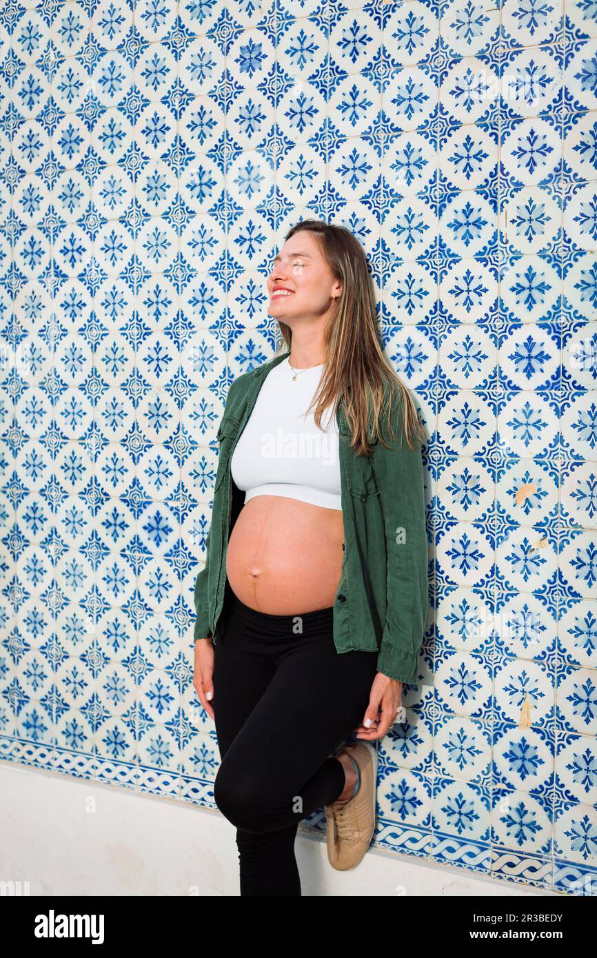 Donna incinta sorridente con gli occhi chiusi appoggiandosi su una parete piastrellata a motivi geometrici Foto Stock