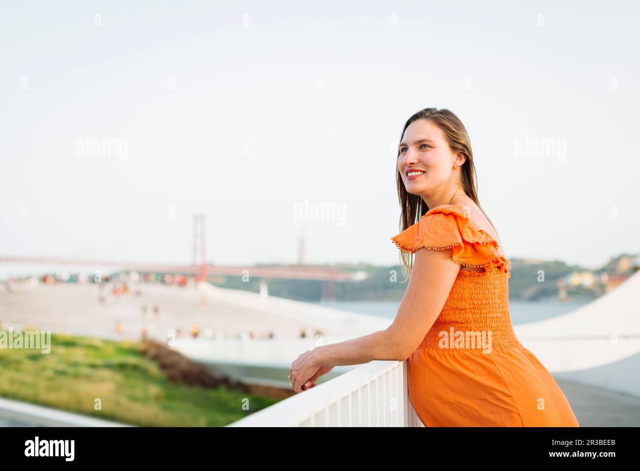 Donna incinta sorridente che indossa un abito arancione appoggiato sulla ringhiera Foto Stock