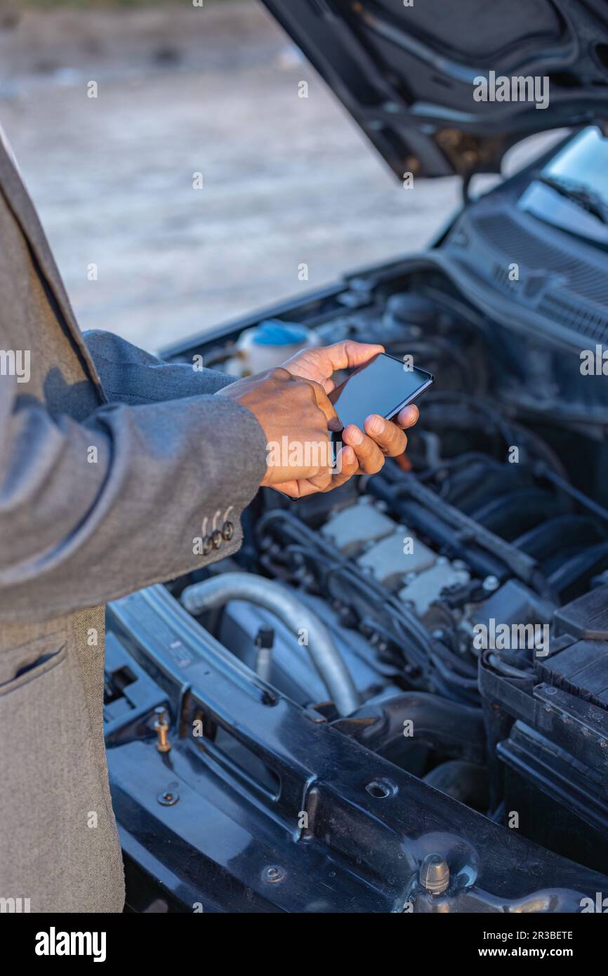 Dettaglio delle mani di un Latino con il suo cellulare con la sua auto rotta sullo sfondo. Foto Stock