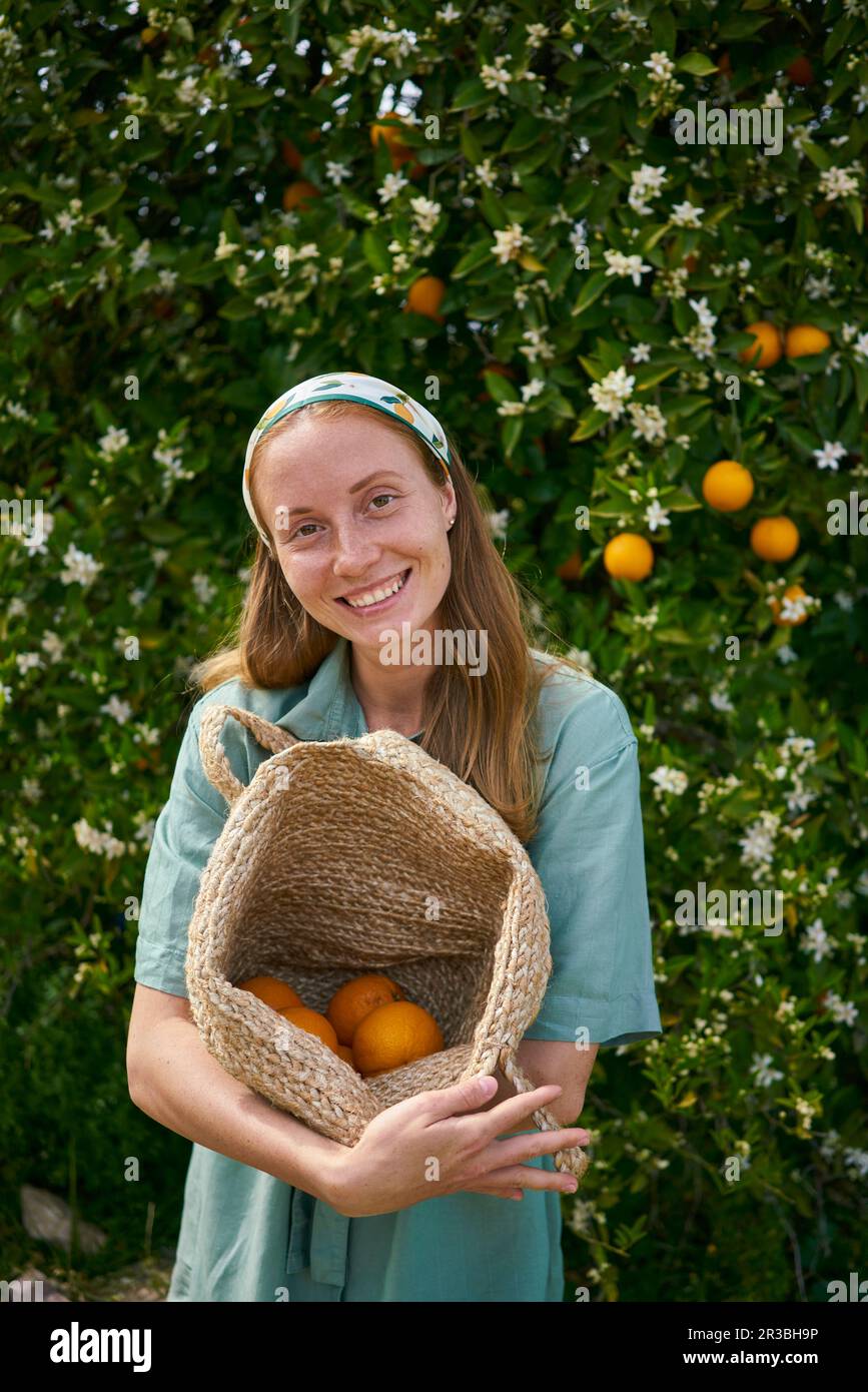 Donna sorridente che tiene un cesto di arance fresche in frutteto Foto Stock