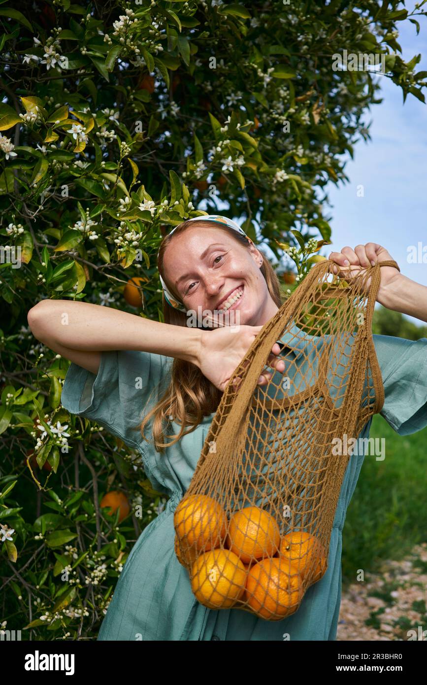 Donna allegra con arance fresche in borsa a rete a frutteto Foto Stock
