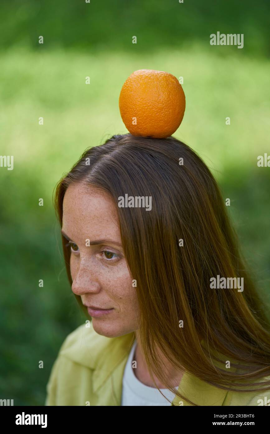 Giovane donna contemplativa equilibrante arancio sulla testa in giardino Foto Stock