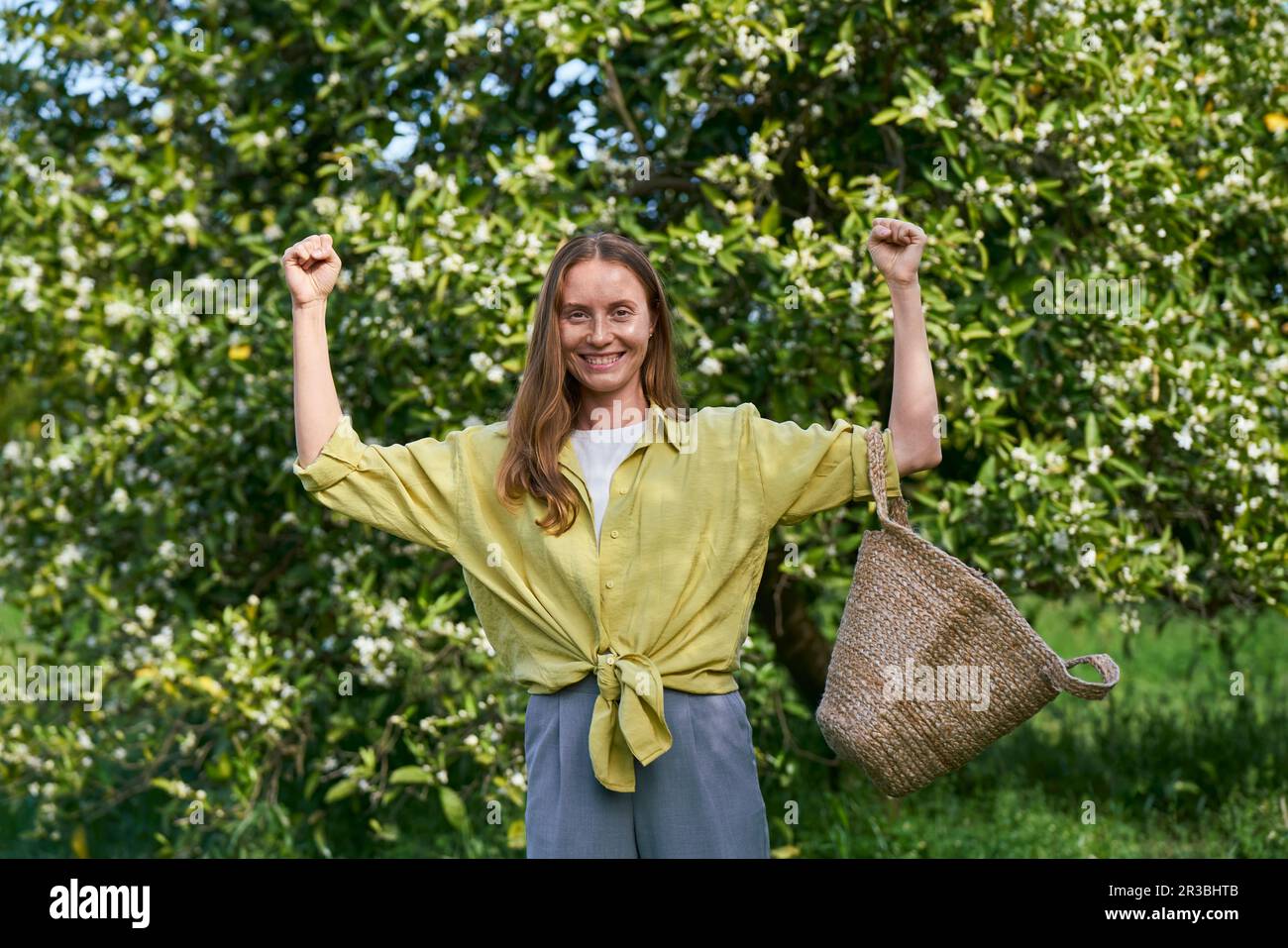 Donna felice che trasporta il sacchetto della paglia in piedi davanti all'albero al frutteto Foto Stock