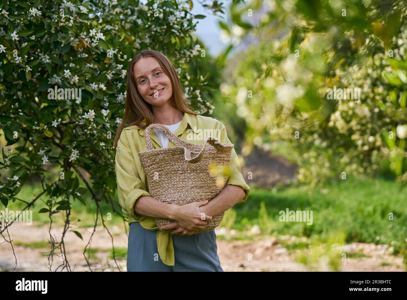 Donna sorridente che tiene la borsa di paglia in piedi in frutteto Foto Stock