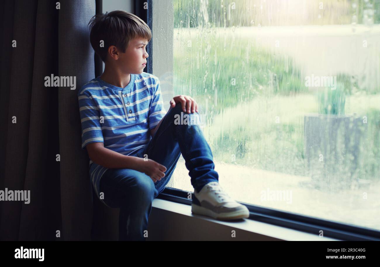 Bambino, a casa e guardando la pioggia dalla finestra di gocce d'acqua naturale e pensando in relax al coperto. Bambino che si rilassa dal davanzale guardando fuori vetro Foto Stock