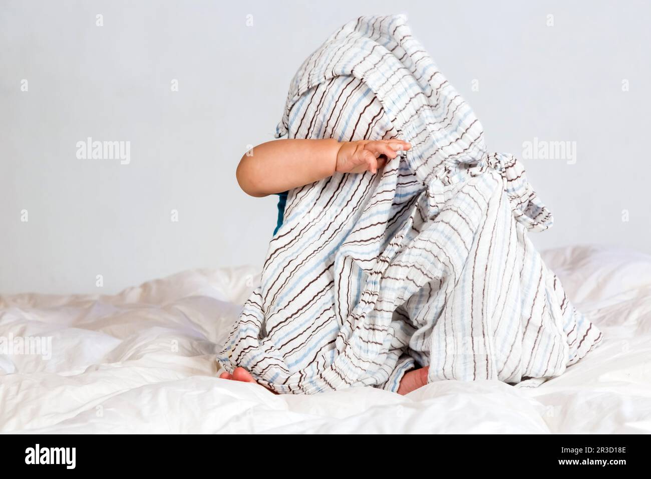 Piccolo ragazzo caucasico che gioca sotto una coperta a strisce su biancheria da letto bianca Foto Stock