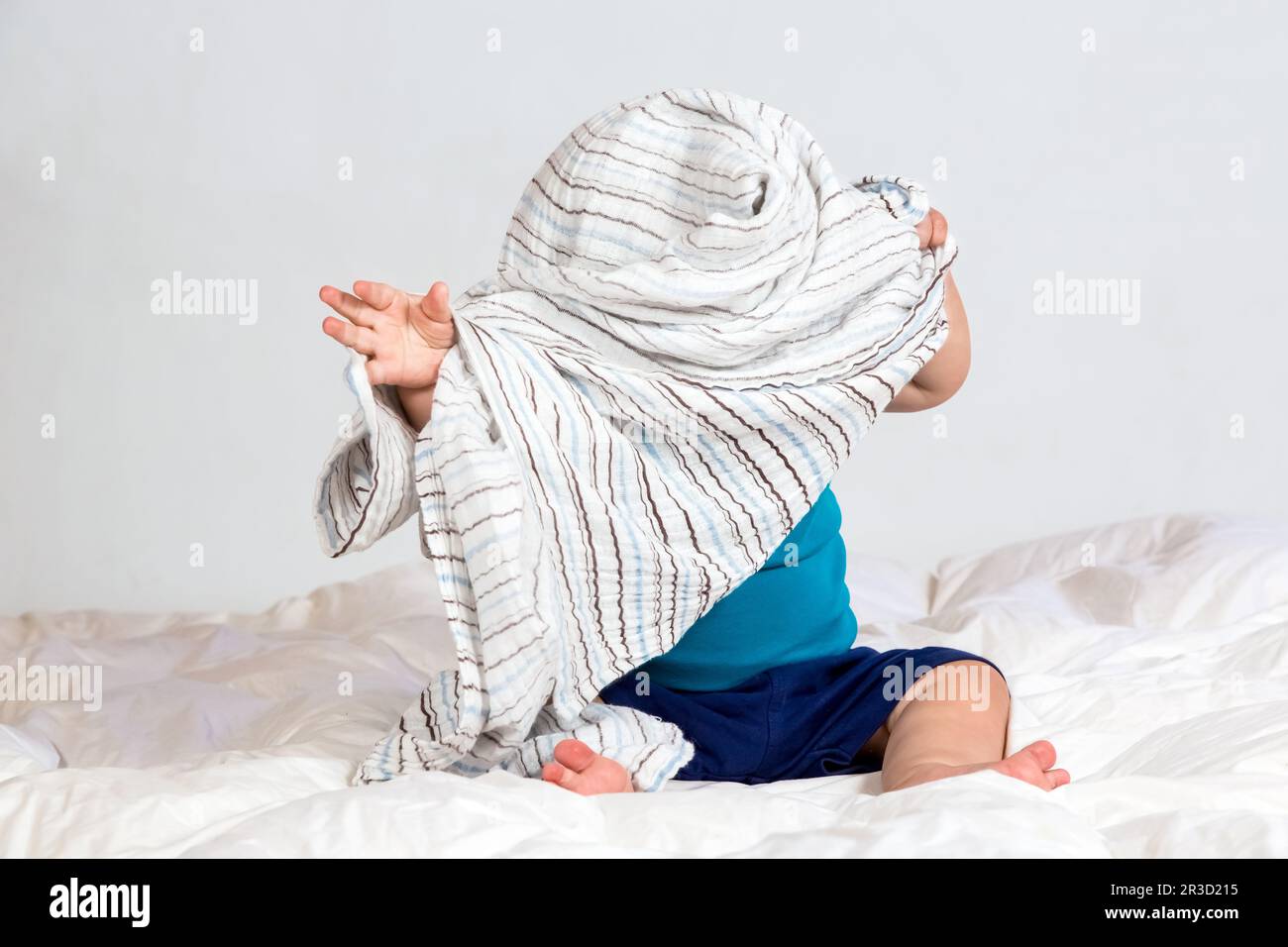Piccolo ragazzo caucasico che gioca sotto una coperta a strisce su biancheria da letto bianca Foto Stock