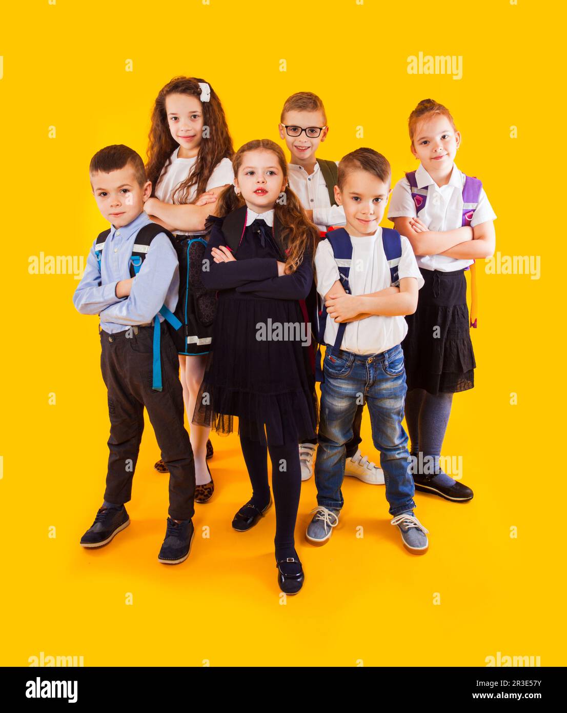 Gruppo di bambini felici in uniforme con borse da scuola abbracciando e guardando alla fotocamera su sfondo giallo Foto Stock