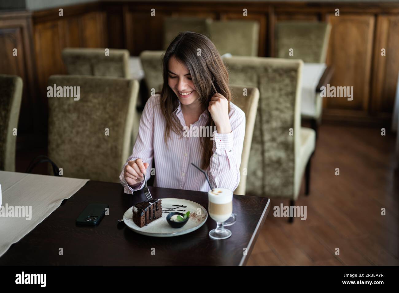 Giovane donna sorridente brunette sedersi al bar caffetteria ristorante al coperto e mangiare torta al cioccolato Brownie dessert. Foto Stock