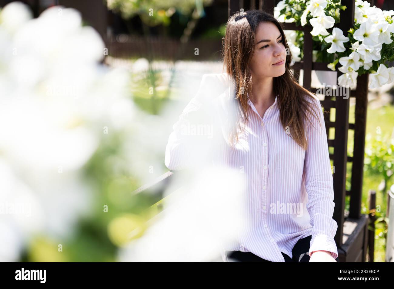 Giovane bella donna in camicia seduta su panca di legno con fiori in giardino. Foto Stock