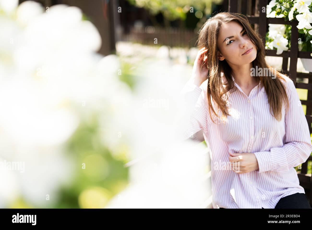Giovane bella donna in camicia seduta su panca di legno con fiori in giardino. Foto Stock