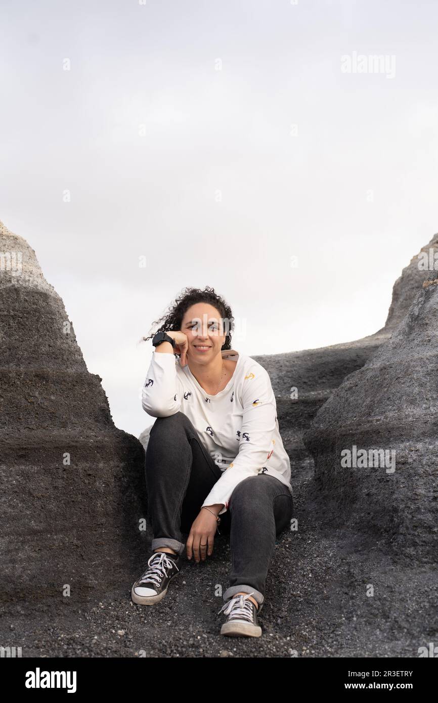 Giovane bruna donna sorridente tra le pietre e le forme della Città stratificata di Lanzarote. Indossa un jumper bianco, jeans neri e scarpe da ginnastica nere. Foto Stock