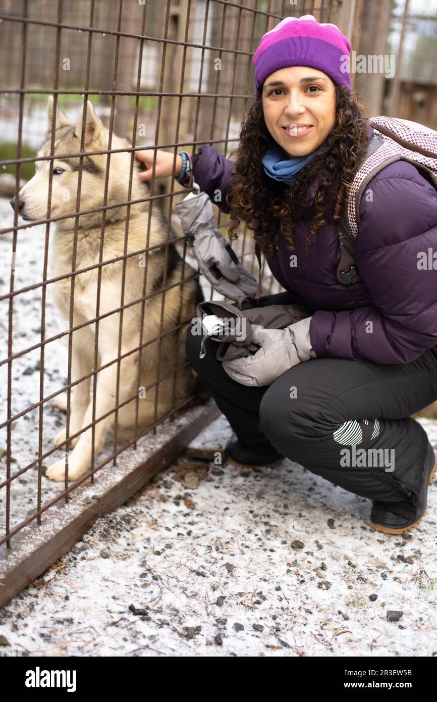 giovane donna brunetta inginocchiata in abiti invernali e cappello accarezzando un cane husky dietro una recinzione Foto Stock