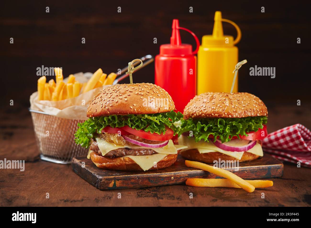 Due hamburger fatti in casa con manzo, formaggio e marmellata di cipolle su un asse di legno, patatine fritte in un cestino di metallo e salse. Conc. Fast food Foto Stock
