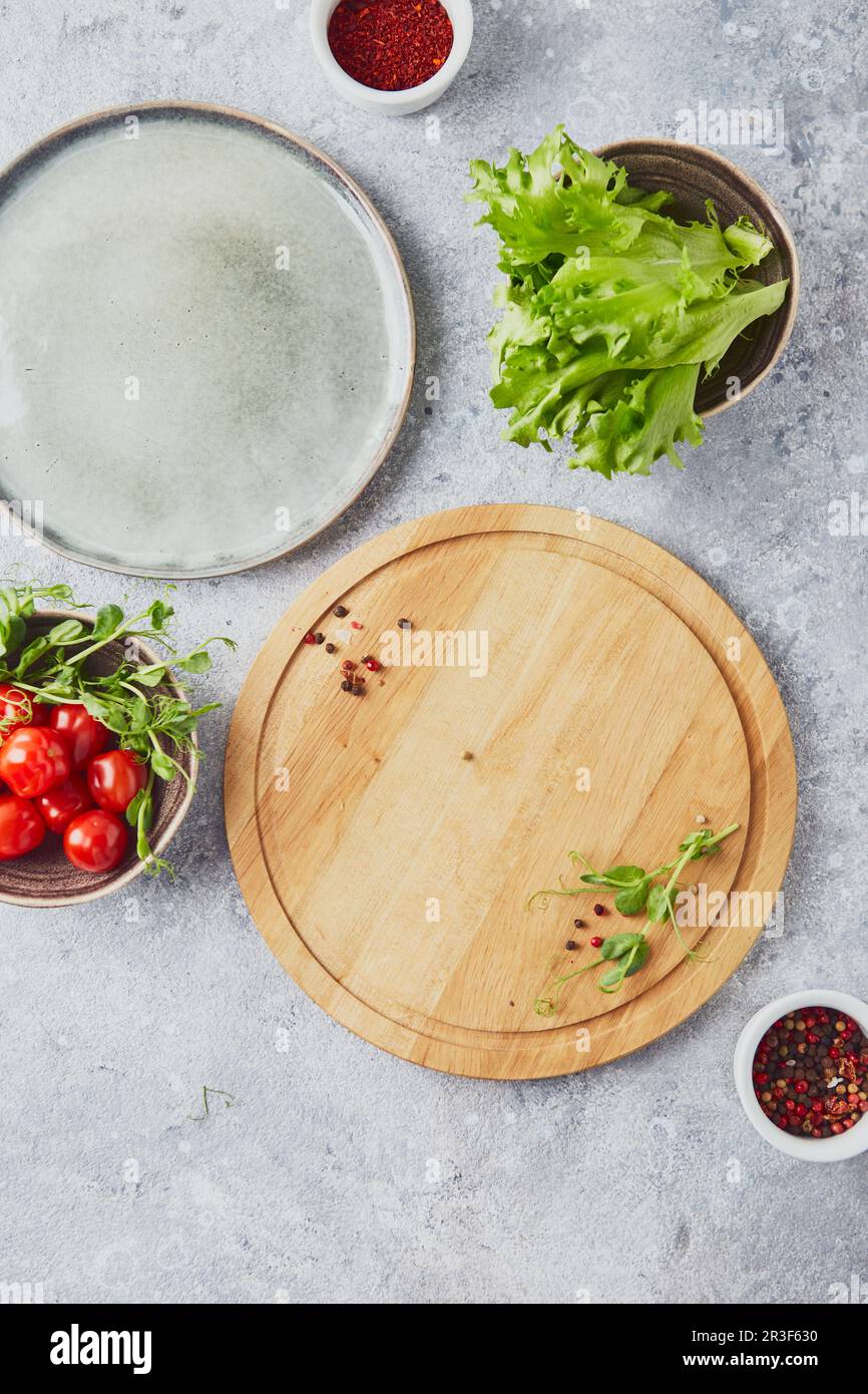 Tagliere rotondo in legno, verdi, verdure e condimenti su sfondo chiaro Foto Stock