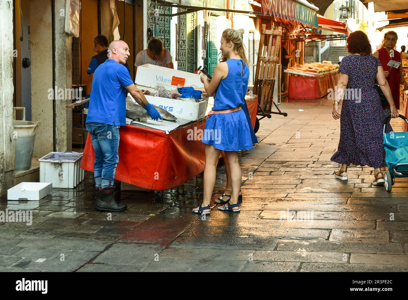 Due giovani turisti stranieri stanno chiacchierando scherzosamente con un pescivendolo che vende pesce gatto al mercato ittico di Rialto in estate, Venezia, Veneto Foto Stock