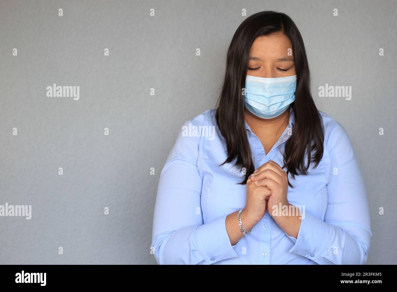 Donna latina con blusa blu e maschera protettiva uso clinico, pregando per pandemia covida Foto Stock