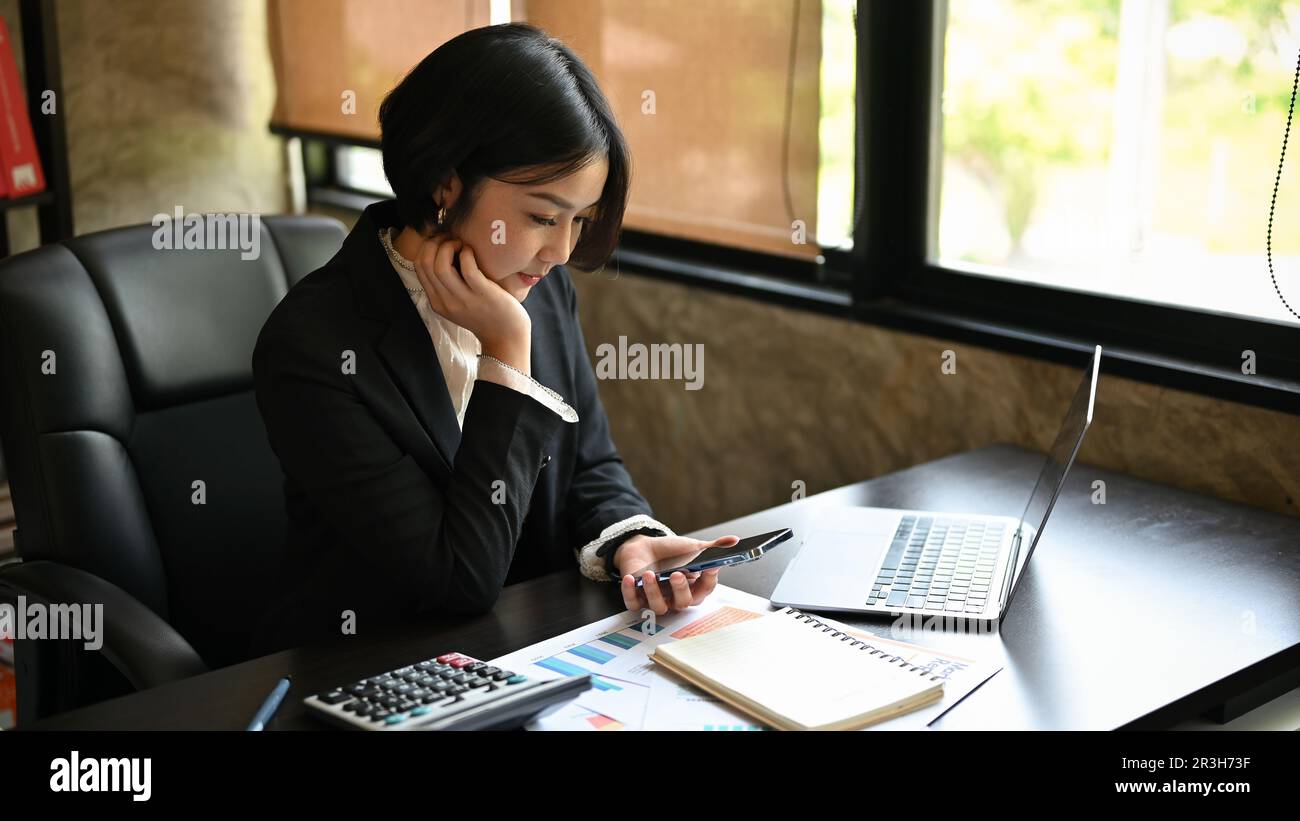 Una donna d'affari asiatica focalizzata mano sul mento, leggendo un articolo online sul sito web attraverso il suo smartphone alla sua scrivania. Foto Stock