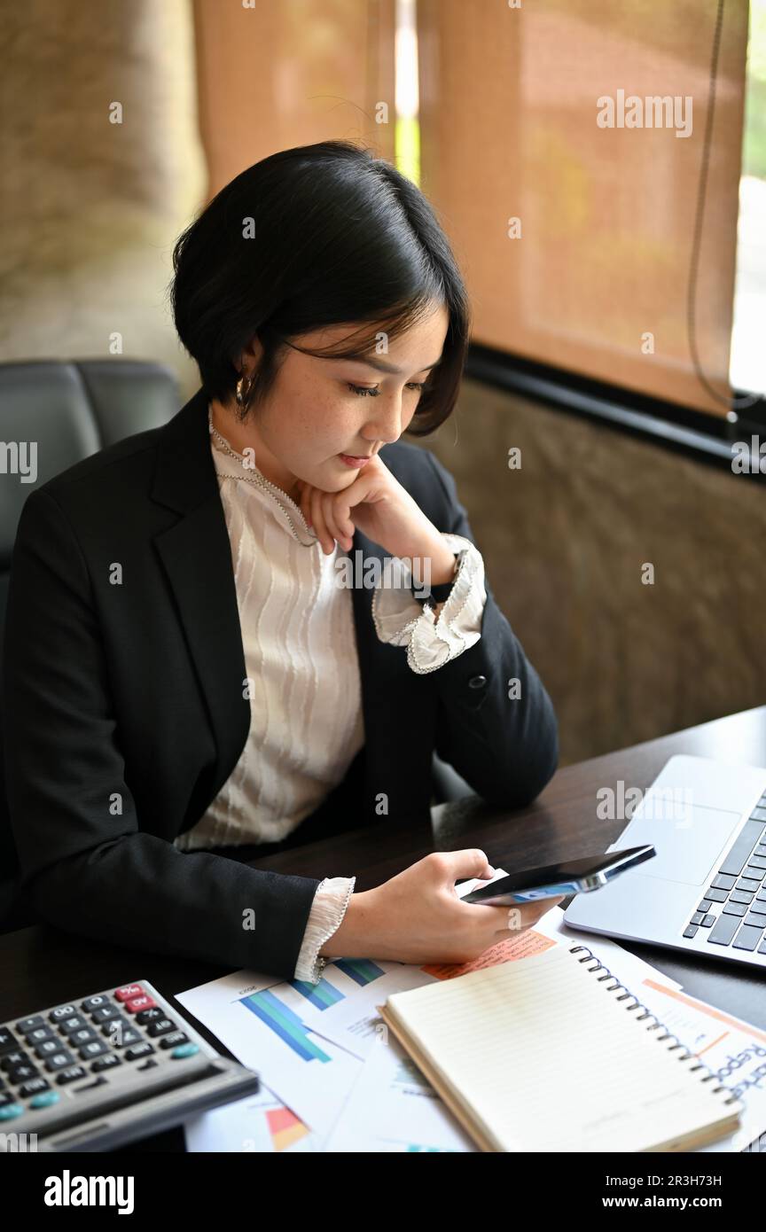 Ritratto di una bella donna d'affari asiatica affascinante controllare i messaggi o leggere un articolo online sul sito web attraverso il suo smartphone alla sua scrivania. Foto Stock