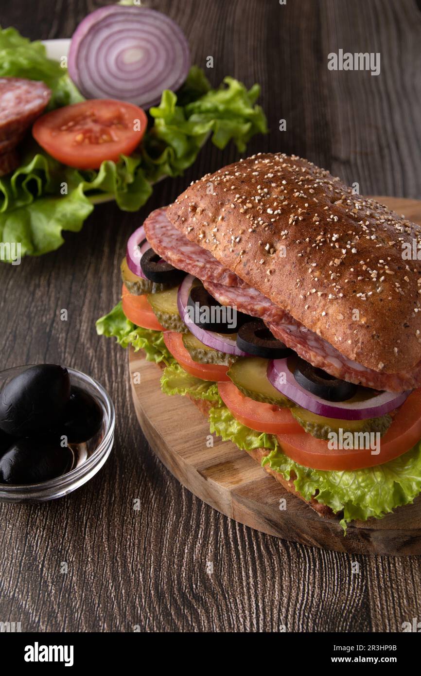 sandwich fotografico con salsiccia, olive, pomodoro, foglie di lattuga e sottaceti su un asse di legno, foto verticale Foto Stock