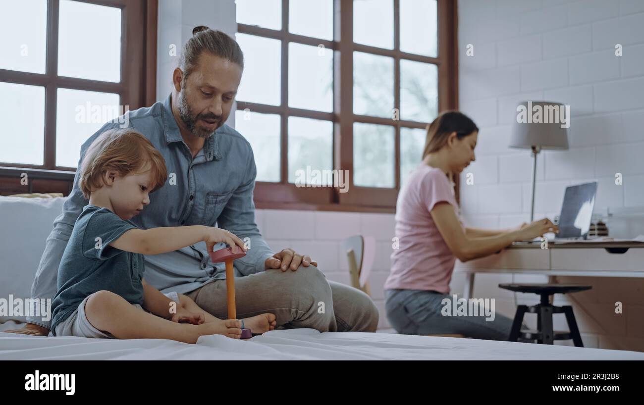 Padre e figlio giocano a giocattolo mentre la madre lavora da casa utilizzando un computer portatile seduto al tavolo. Foto Stock