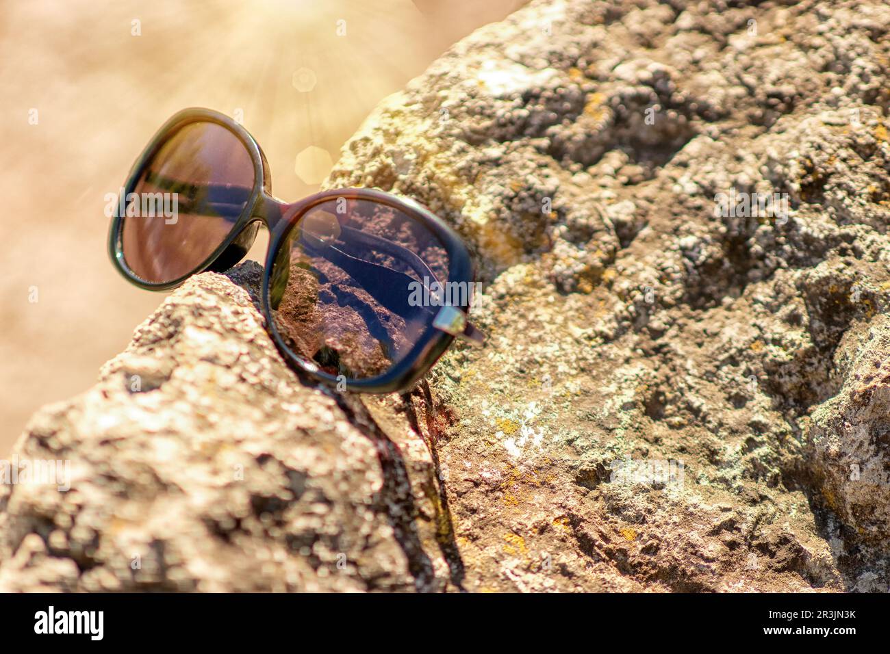 gli occhiali da sole giacciono su una pietra testurizzata, sulla riva del mare, sotto i raggi diretti del sole bruciante, il riflesso dei raggi del sole si riflette sugli occhiali Foto Stock