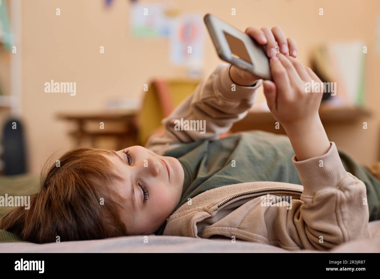 Ritratto di carino ragazzo tenendo smartphone mentre sdraiato sul letto e sorridente, gen Alpha Foto Stock