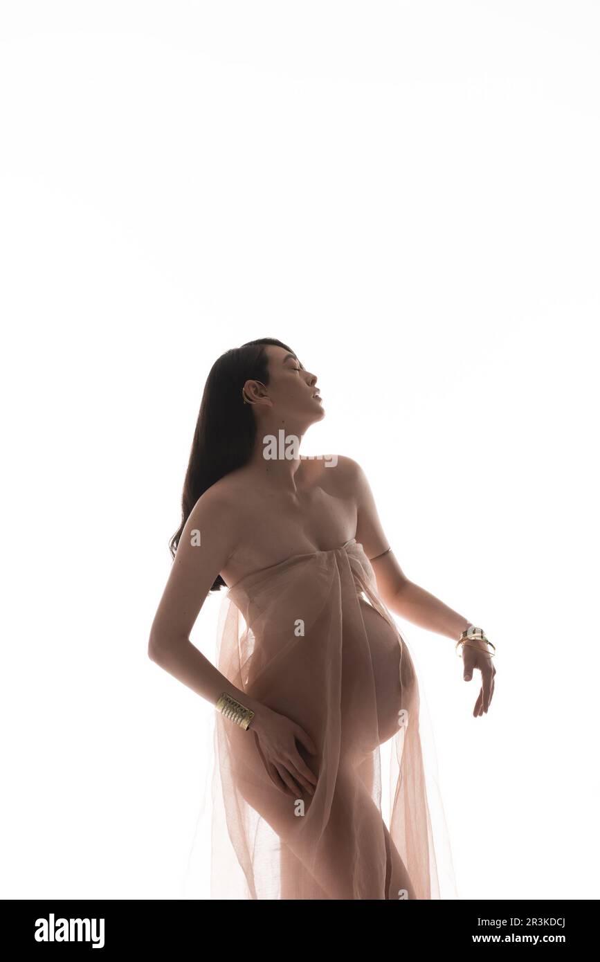 affascinante e romantica donna incinta posa in accessori dorati e trasparente chiffon drappeggio isolato su sfondo bianco, conc. moda maternità Foto Stock