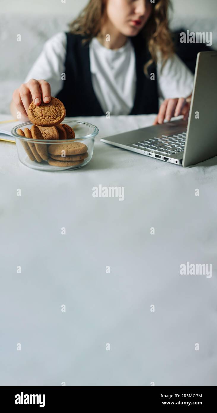 Mangiare inconscio. Cattive abitudini. Fuoco selettivo della mano di donne concentrate che lavorano sul laptop al tavolo della cucina, prendendo involontariamente e. Foto Stock