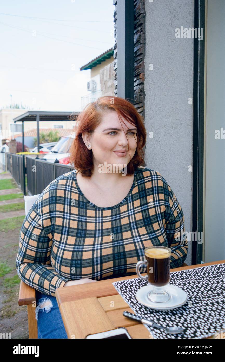 giovane donna latina di più dimensioni con capelli rossi è seduta fuori dalla caffetteria sorridendo guardando dritto in avanti, prestando attenzione alla chat del suo amico. vert Foto Stock