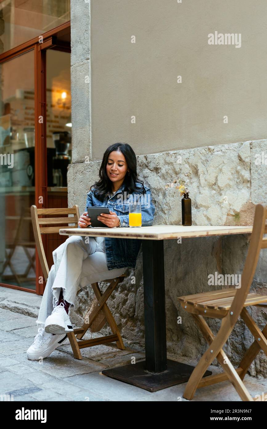Ragazza Happy Hispanic in giacca denim seduta a un tavolo da caffè croce zampe tenendo il suo tablet durante la navigazione in rete Foto Stock