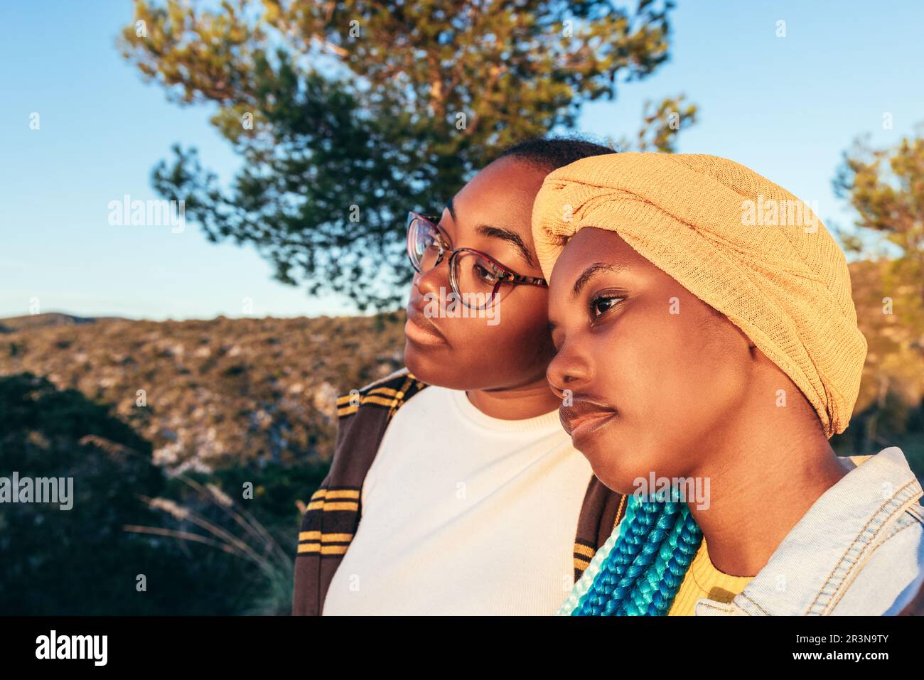 Da sotto di giovani donne afro-americane premurose in copricapo e occhiali che guardano via mentre si levano in piedi contro l'albero verde e blu senza nuvole s Foto Stock