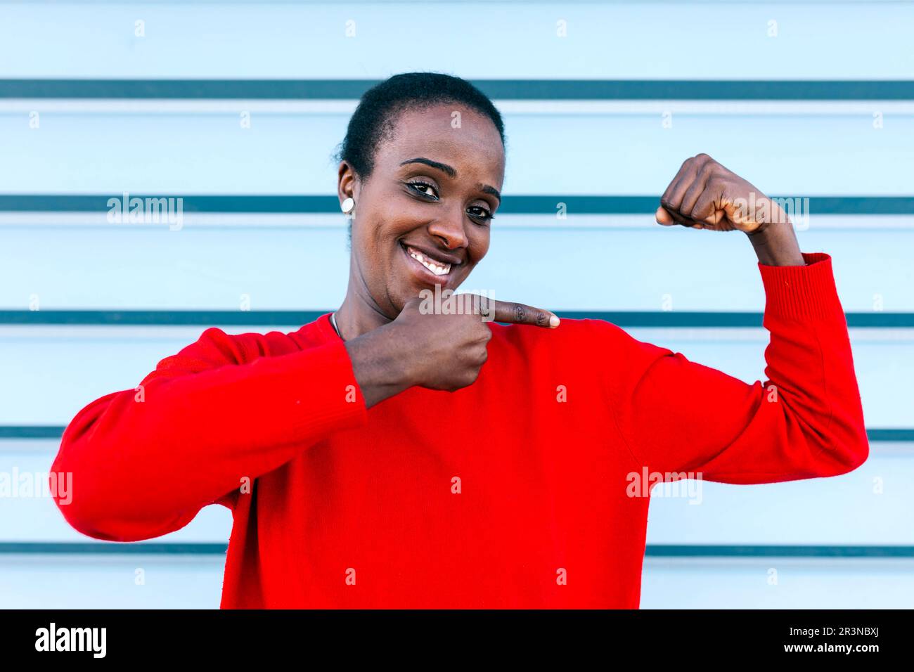 Ritratto di contenuti modello afro-americano femminile guardando la fotocamera e sorridendo mentre indica il bicep e in piedi contro il muro blu in città Foto Stock