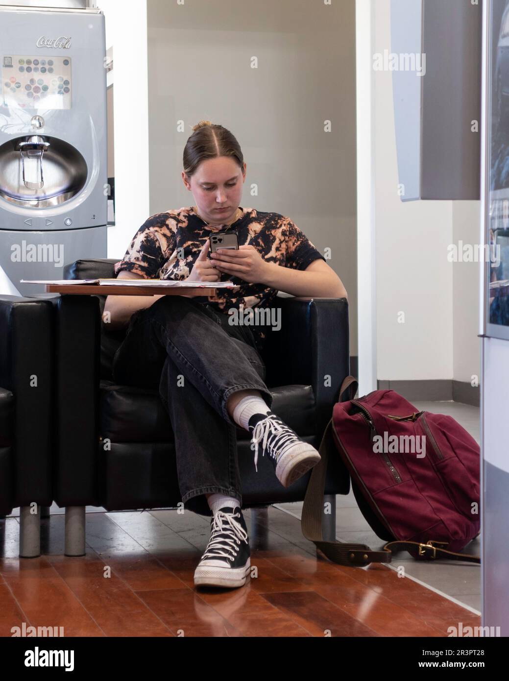 Una giovane donna caucasica con uno zaino rosso in una sala d'attesa di servizio auto guardando il suo cellulare. Kansas, Stati Uniti. Foto Stock