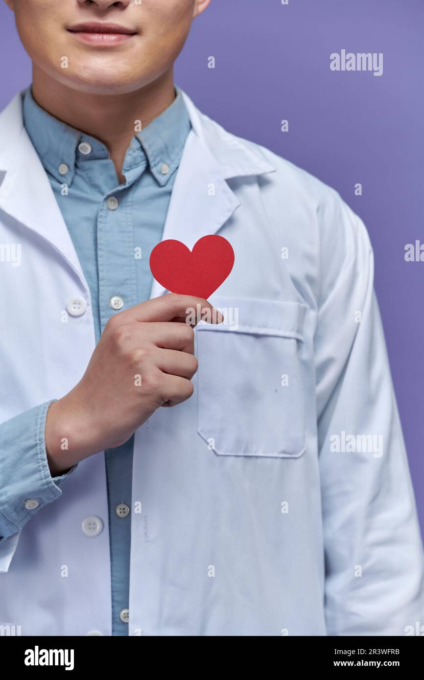 Giovane medico con stetoscopio su sfondo viola tenendo il cuore rosso Foto Stock