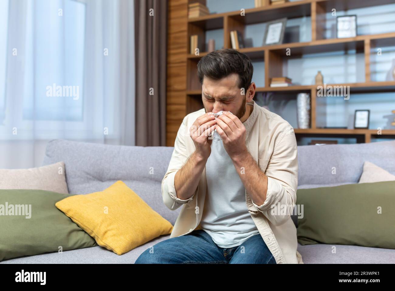 Malato uomo maturo seduto sul divano a casa, l'uomo ha allergia e naso che cola, starnuti in salotto da solo. Foto Stock