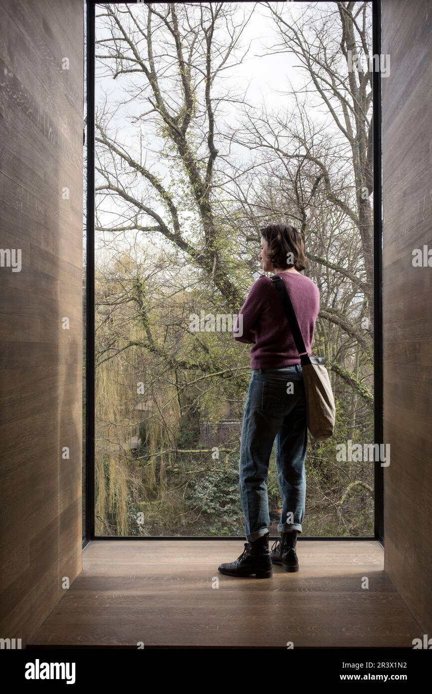 Una vista posteriore di una donna in jeans e un maglione viola come lei si leva in piedi in una stanza rivestita di legno che guarda da una grande finestra di vetro di lastra a livello dell'albero. Foto Stock