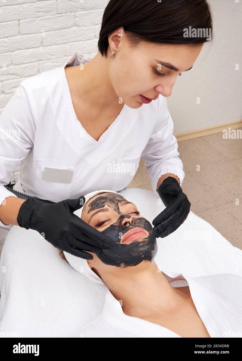 Cosmetologo applicazione maschera nera sul viso di una bella donna per buccia di carbonio Foto Stock