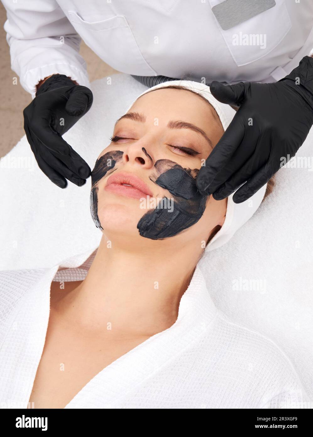 Cosmetologo applicazione maschera nera sul viso di una bella donna per buccia di carbonio Foto Stock