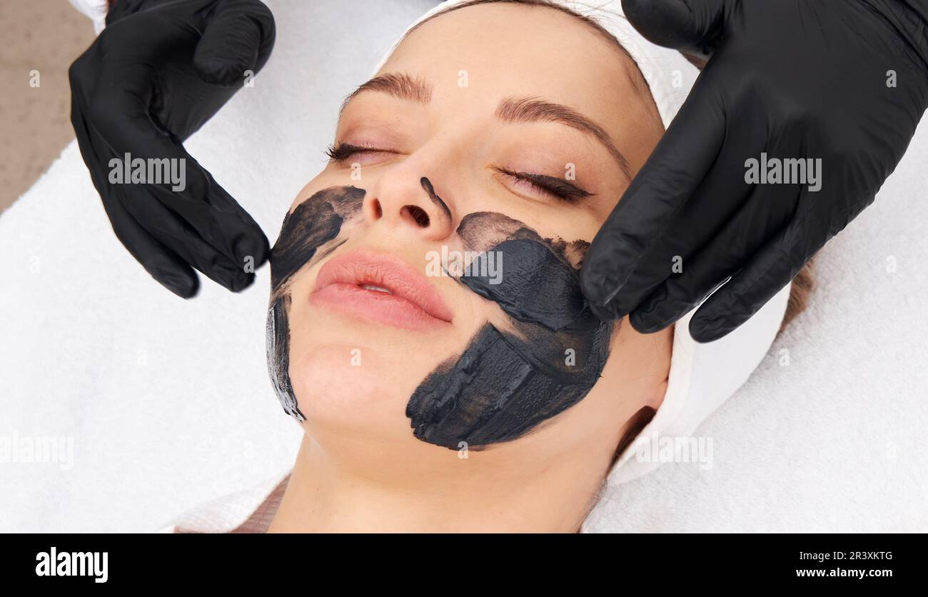 Cosmetologo applicazione maschera nera sul viso di una bella donna per buccia di carbonio Foto Stock