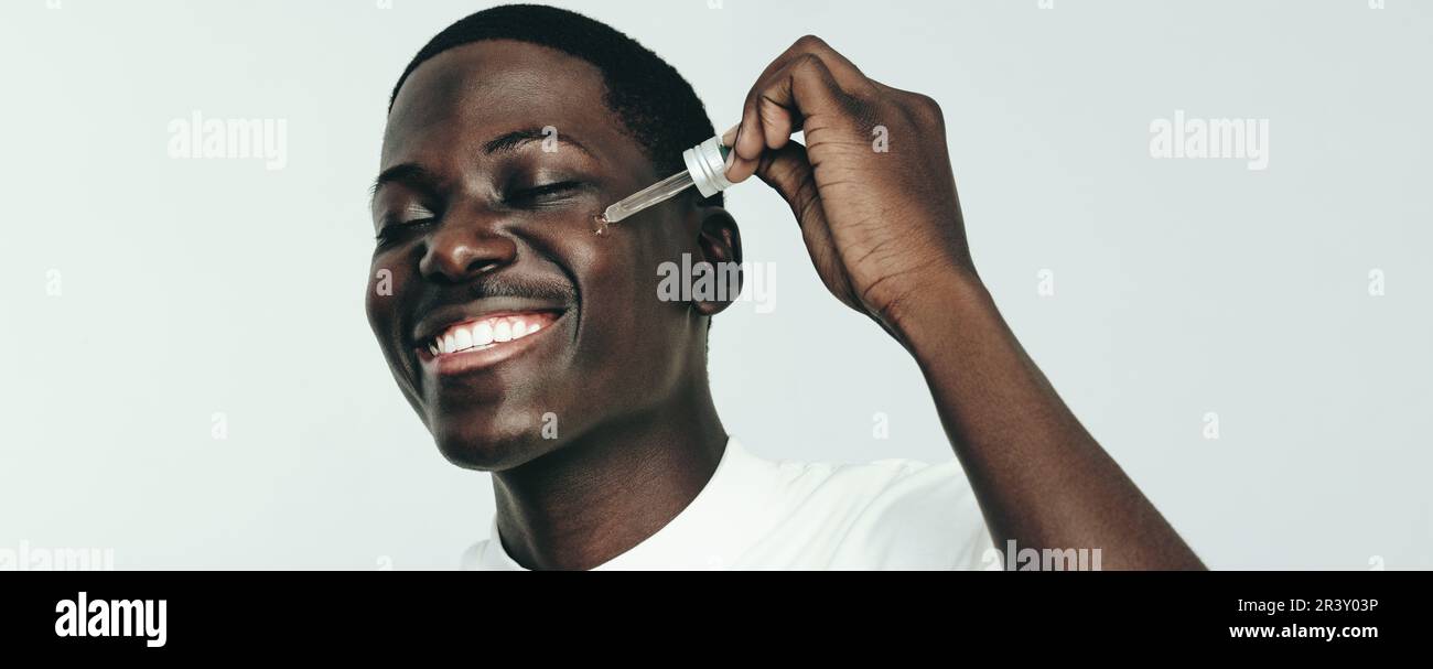 Giovane uomo nero sorridente mentre applica olio di bellezza al suo viso melanated in uno studio. Felice uomo giovane con orgoglio praticare la sua routine di cura della pelle con un Foto Stock