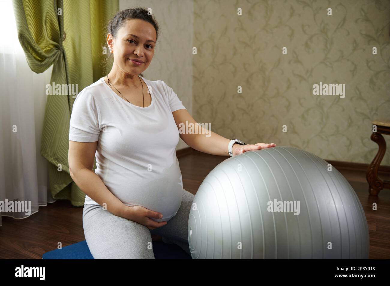 Autentica donna incinta sorride alla macchina fotografica, tocca il ventre, seduto su un tappetino yoga mentre si esercita con la palla fit Foto Stock