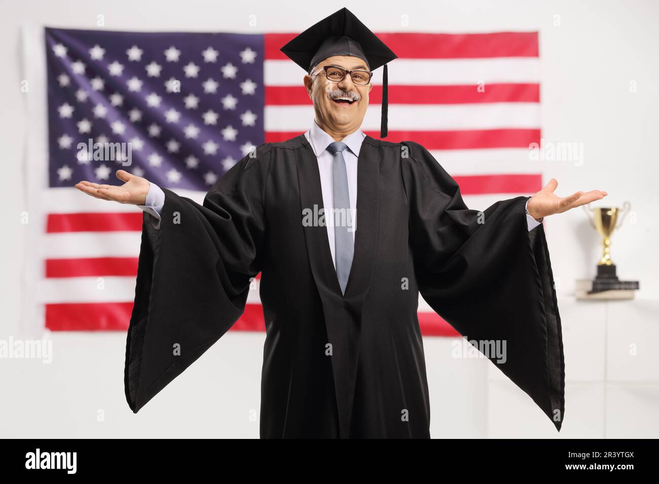 Uomo maturo allegro che indossa un abito da laurea di fronte alla bandiera degli Stati Uniti Foto Stock