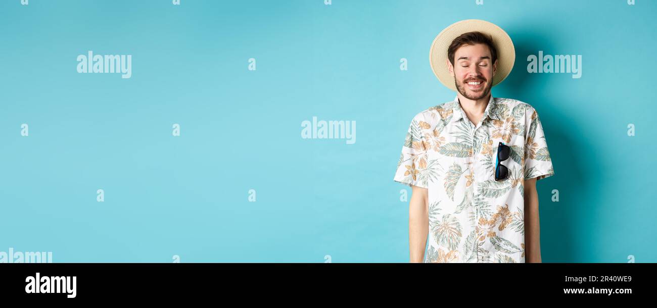 Felice turista godendo di vacanza estiva, con cappello di paglia e camicia hawaiana, in piedi gioioso su sfondo blu Foto Stock