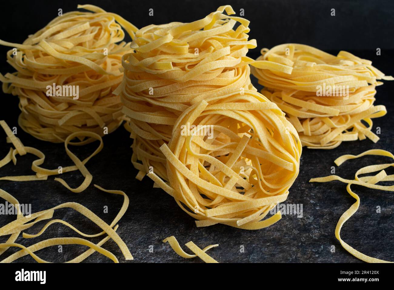 Nidi di tagliatelle impilati e noodle sparsi su sfondo scuro: Vista laterale e dall'alto di nidi di pasta essiccata impilati sopra l'altro Foto Stock