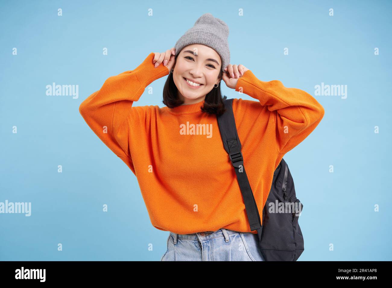 Ragazza asiatica sorridente carina mette su cappello caldo per andare fuori, passeggiate con zaino in maglione arancione, sfondo blu Foto Stock