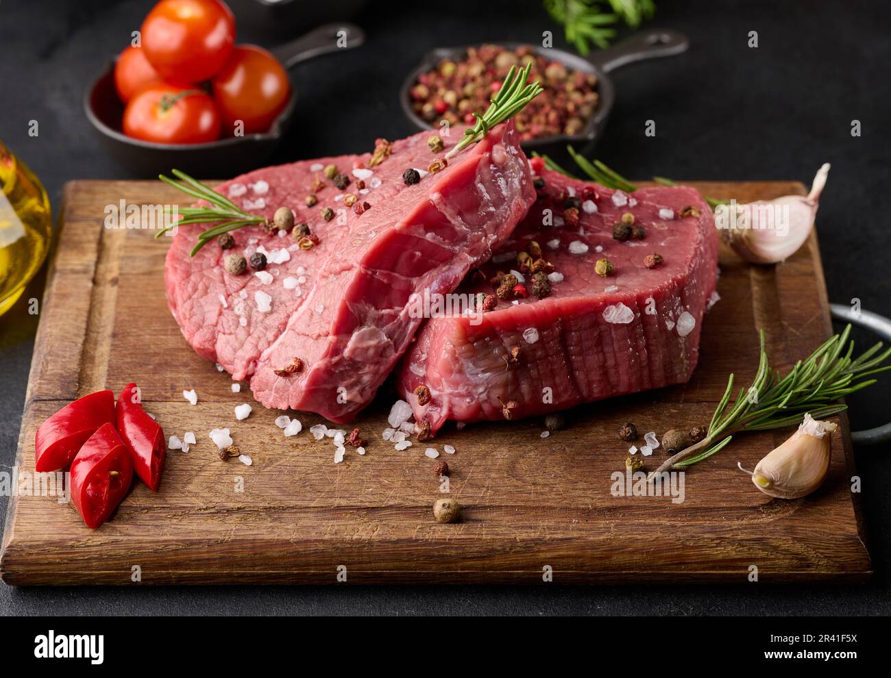 Pezzo di manzo crudo con spezie, pepe, rametto di rosmarino, sale e olio d'oliva su un asse di legno, fondo nero Foto Stock